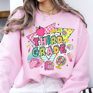 K&ouml;nnte beinhalten: Rosafarbener Pullover mit einem Grafikdesign aus einem Regenbogen, einem Globus, einem Rucksack, einem Bleistift, einem Buch, einem roten Apfel und dem Text "Third Grade".