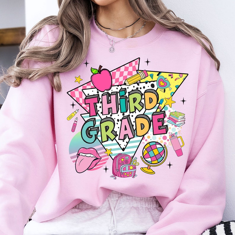 K&ouml;nnte beinhalten: Rosafarbener Pullover mit einem Grafikdesign aus einem Regenbogen, einem Globus, einem Rucksack, einem Bleistift, einem Buch, einem roten Apfel und dem Text "Third Grade".