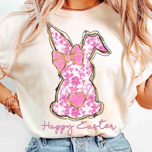 Op de afbeelding: Een wit T-shirt met een roze bloemachtig konijnenontwerp en de tekst "Happy Easter" gedrukt op de voorkant.