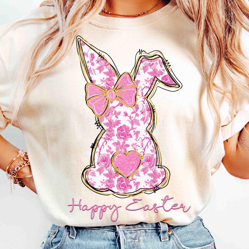 Op de afbeelding: Een wit T-shirt met een roze bloemachtig konijnenontwerp en de tekst "Happy Easter" gedrukt op de voorkant.