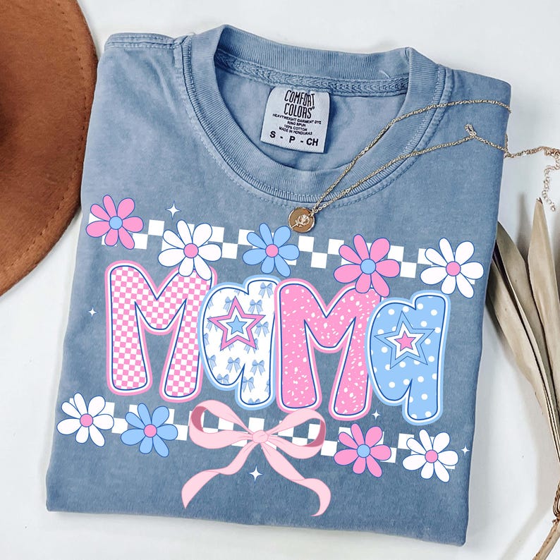 May include: A blue Comfort Colors t-shirt with the word "MAMA" in pink and blue, decorated with flowers and a bow. The shirt has a round neckline and short sleeves. A gold necklace is draped across the shirt.