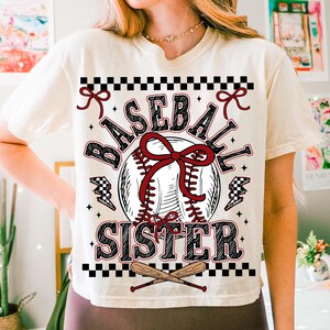 May include: Cream-colored t-shirt with a baseball-themed graphic. The design features a baseball with a red bow, the words "BASEBALL SISTER", and crossed bats. Accents include black and white checkered patterns and lightning bolts.