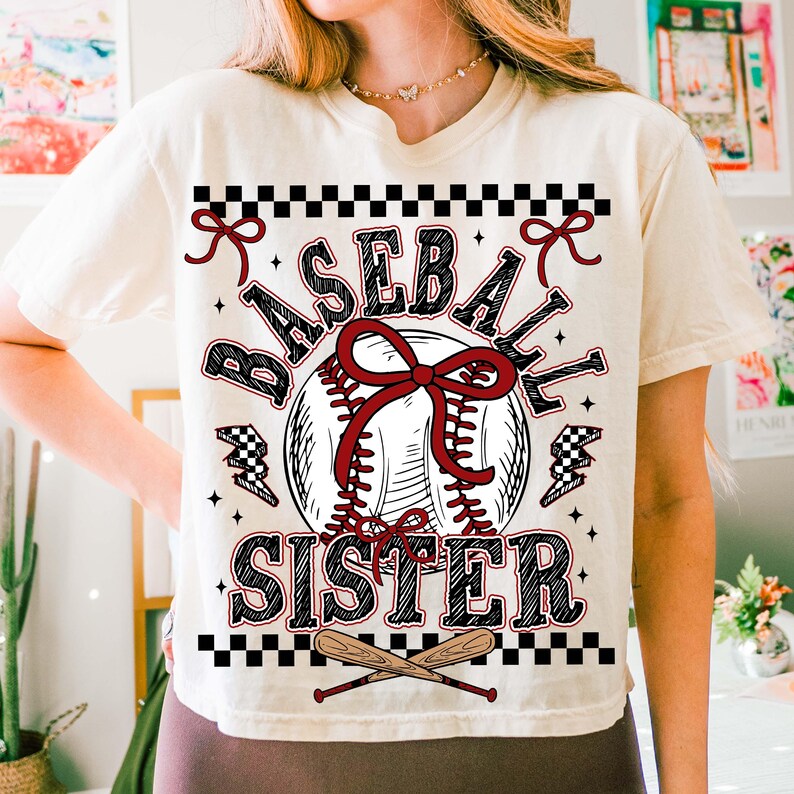 May include: Cream-colored t-shirt with a baseball-themed graphic. The design features a baseball with a red bow, the words "BASEBALL SISTER", and crossed bats. Accents include black and white checkered patterns and lightning bolts.