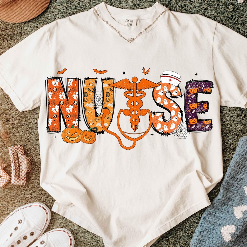 May include: Cream-colored t-shirt with a Halloween-themed design. The word "NURSE" is spelled out in orange and purple letters with Halloween graphics. A stethoscope and medical symbol are also featured in orange. Bats, pumpkins, and spiderwebs complete the design.