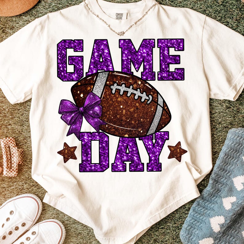 Puede incluir: Camiseta color crema con un dise&ntilde;o de purpurina morada "GAME DAY". El dise&ntilde;o presenta un bal&oacute;n de f&uacute;tbol americano marr&oacute;n con rayas plateadas y un lazo morado. Dos estrellas marrones flanquean la palabra "DAY".
