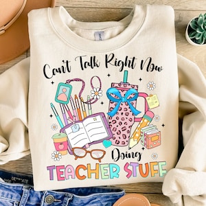 May include: A beige sweatshirt with the text "Can't Talk Right Now Doing Teacher Stuff" in rainbow colors. The text is surrounded by a variety of school supplies, including a coffee cup, a pencil, a notebook, and a pair of glasses.