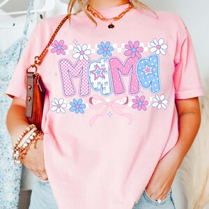 May include: Pink t-shirt with the word "MAMA" in a retro font. The letters are decorated with checkered patterns, stars, and polka dots. Floral and bow accents surround the text, creating a cheerful and feminine design.