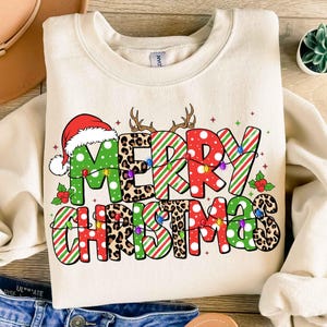 May include: A beige sweatshirt with a Christmas design. The design features the words "Merry Christmas" in green, red, and white stripes, leopard print, and white polka dots. The letters are decorated with red and green lights.
