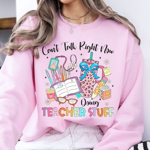 May include: Pink sweatshirt with the text "Can't Talk Right Now Doing Teacher Stuff" and a graphic of a coffee cup, books, pencils, and glasses.