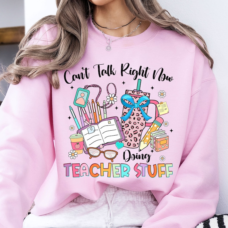 May include: Pink sweatshirt with the text "Can't Talk Right Now Doing Teacher Stuff" and a graphic of a coffee cup, books, pencils, and glasses.