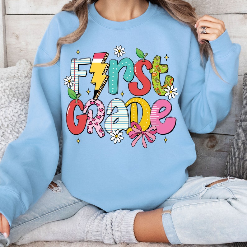 May include: A light blue sweatshirt with the text "First Grade" in colorful, playful letters. The letters are decorated with various school-themed patterns, including a lightning bolt, a pencil, and a heart.