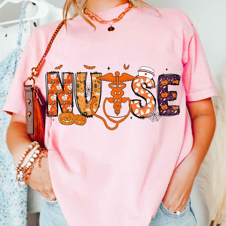 May include: A light pink t-shirt with the word "NURSE" in a Halloween-themed design. The letters are decorated with pumpkins, ghosts, and spiderwebs. A stethoscope graphic is included in the design.