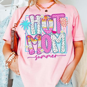 Puede incluir: Camiseta rosa con un gr&aacute;fico colorido que dice "HOT MOM summer". El dise&ntilde;o incluye una palmera, flores, un sol y una rodaja de sand&iacute;a. Las letras son blancas con lunares negros y patrones de cuadros rosas.