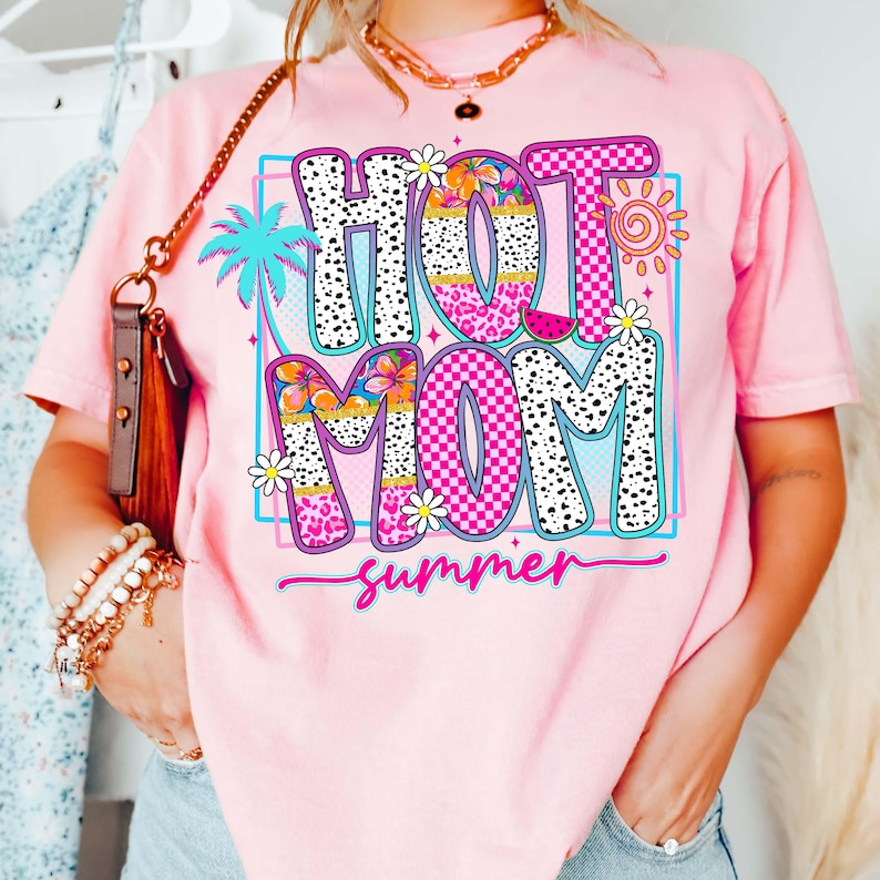 Puede incluir: Camiseta rosa con un gr&aacute;fico colorido que dice "HOT MOM summer". El dise&ntilde;o incluye una palmera, flores, un sol y una rodaja de sand&iacute;a. Las letras son blancas con lunares negros y patrones de cuadros rosas.