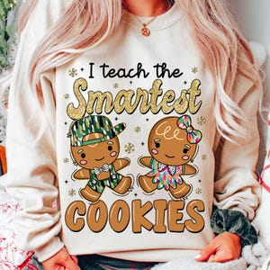 May include: A white sweatshirt with the text "I teach the smartest cookies" in gold glitter lettering. The design features two gingerbread people, one wearing a green and brown camouflage hat and the other wearing a pink and green bow.