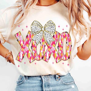 K&ouml;nnte beinhalten: Cremefarbenes Sweatshirt mit den Buchstaben "XOXO" in rosa, gold- und wei&szlig; gemustert, mit einer schwarz-wei&szlig; gepunkteten Schleife. Rosa Sternakzente umgeben den Text und schaffen ein verspieltes Design.