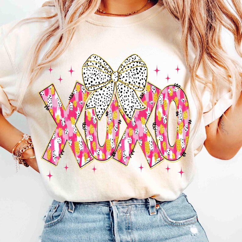 K&ouml;nnte beinhalten: Cremefarbenes Sweatshirt mit den Buchstaben "XOXO" in rosa, gold- und wei&szlig; gemustert, mit einer schwarz-wei&szlig; gepunkteten Schleife. Rosa Sternakzente umgeben den Text und schaffen ein verspieltes Design.