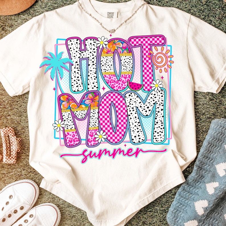 Puede incluir: Camiseta color crema con un gr&aacute;fico colorido que dice "HOT MOM summer". El dise&ntilde;o presenta una palmera, flores, un sol y una rodaja de sand&iacute;a. Las letras son rosas, negras y blancas.
