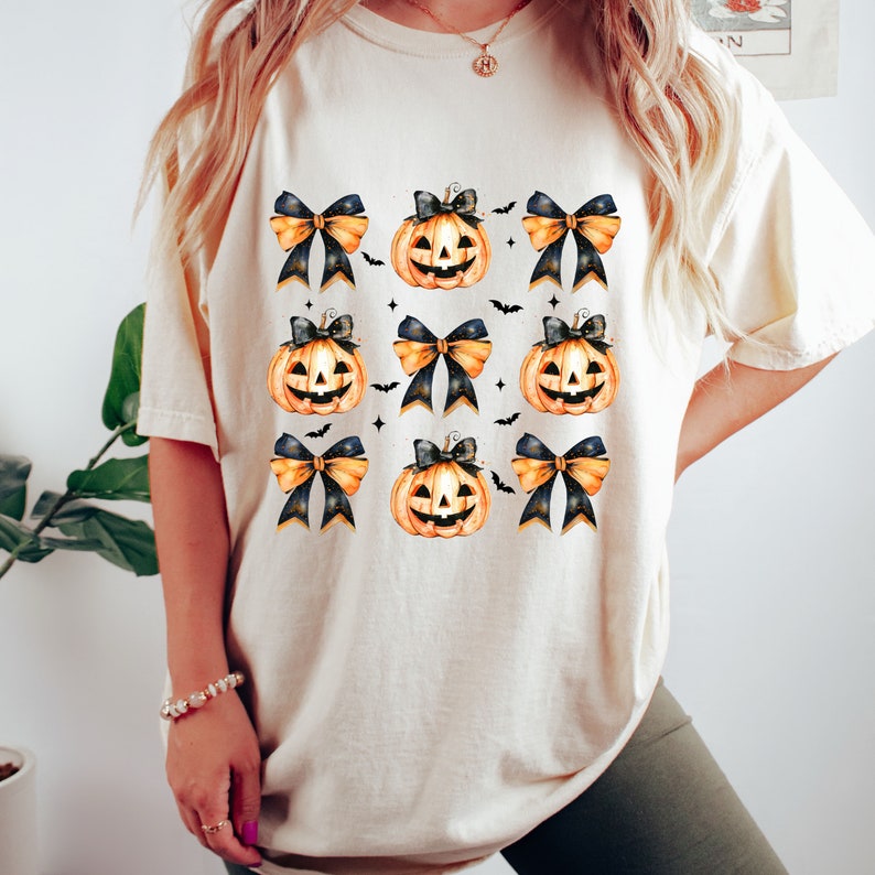 May include: A white t-shirt with a pattern of orange jack-o'-lantern pumpkins with black and gold bows. The pumpkins have black eyes, a black nose, and a wide, toothy grin.