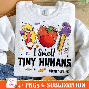 May include: Light gray sweatshirt with a colorful graphic design. The design includes a pencil, apple, and ruler with the text "I Smell TINY HUMANS" and "#TeacherLife". The sweatshirt is a Gildan brand.