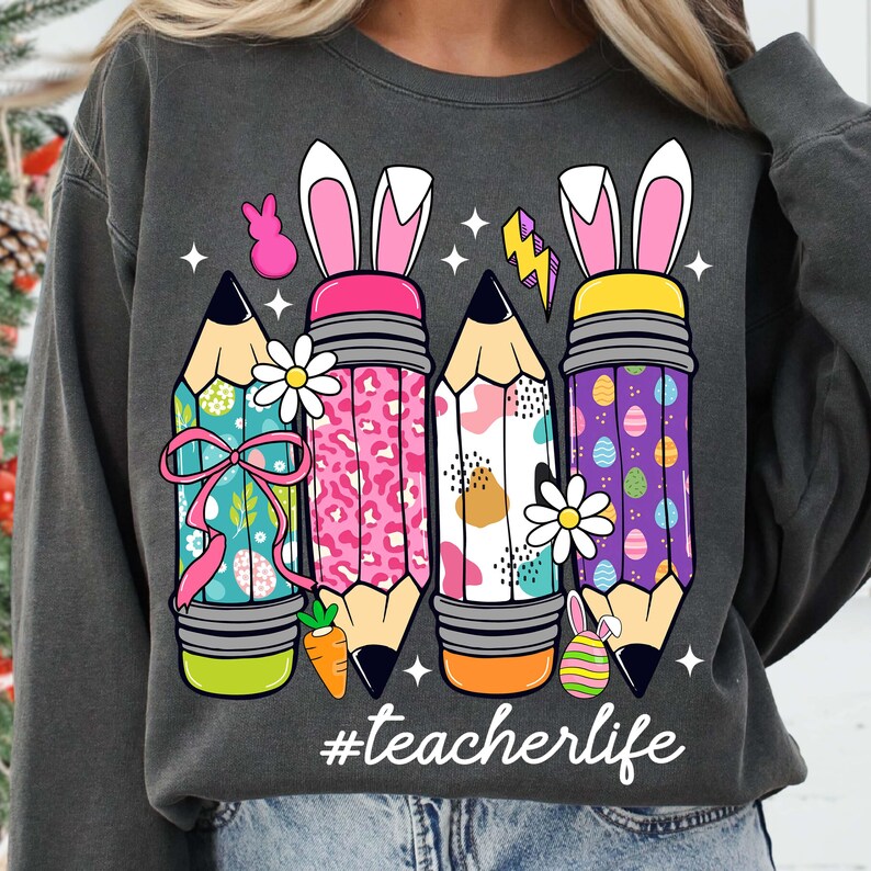 May include: A grey sweatshirt featuring a graphic of four pencils with bunny ears and Easter-themed designs. The pencils are decorated with polka dots, animal print, and Easter eggs. The text "#teacherlife" is printed below the pencils.