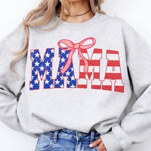 May include: Gray sweatshirt with the word "MAMA" in red, white, and blue glitter letters, with a red, white, and blue American flag pattern. A pink bow is above the word.
