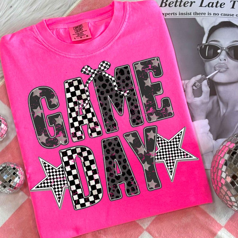 May include: Bright pink t-shirt with the words "GAME DAY" in a bold, playful font. The letters feature a mix of patterns, including stars, leopard print, and checkered designs. A checkered bow adds a decorative touch. The shirt is folded on a pink and white checkered surface.