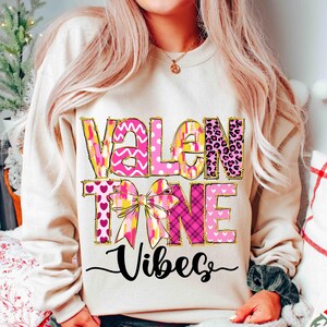 K&ouml;nnte beinhalten: Ein cremefarbenes Sweatshirt mit den Worten "VALENTINE VIBES" in rosa, gelben und goldenen Buchstaben. Das Design umfasst eine Schleife und verschiedene Muster, darunter Herzen und Leopardenmuster. Das Wort "Vibes" ist kursiv geschrieben.