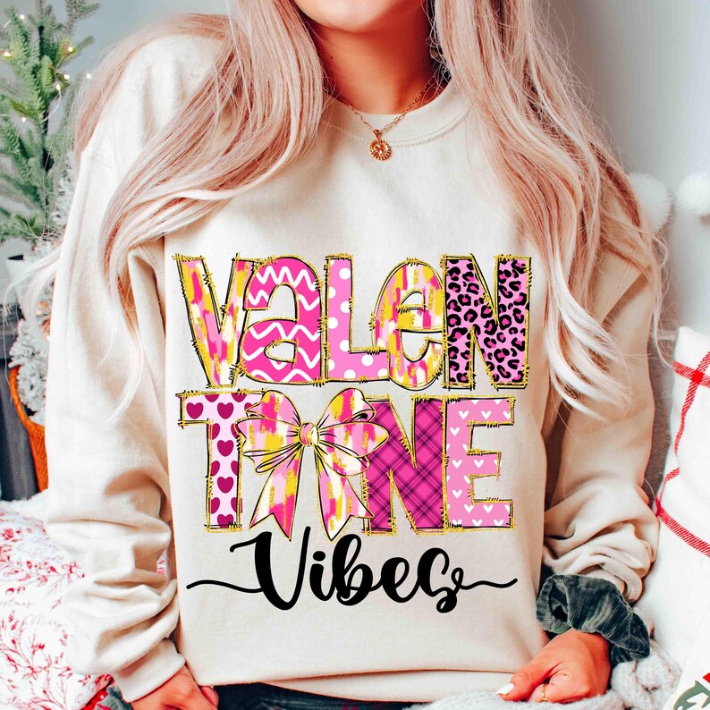 K&ouml;nnte beinhalten: Ein cremefarbenes Sweatshirt mit den Worten "VALENTINE VIBES" in rosa, gelben und goldenen Buchstaben. Das Design umfasst eine Schleife und verschiedene Muster, darunter Herzen und Leopardenmuster. Das Wort "Vibes" ist kursiv geschrieben.