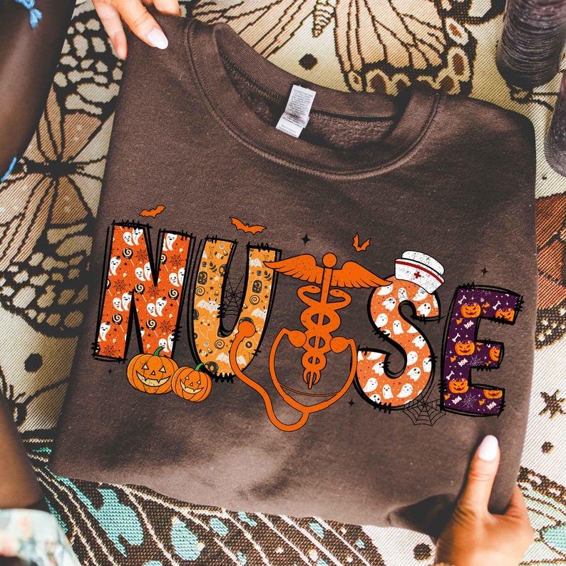 May include: A brown sweatshirt with the word "NURSE" in orange, white, and purple, decorated with Halloween-themed graphics. The "U" features a medical symbol and stethoscope. Pumpkins, ghosts, and bats are also included in the design.