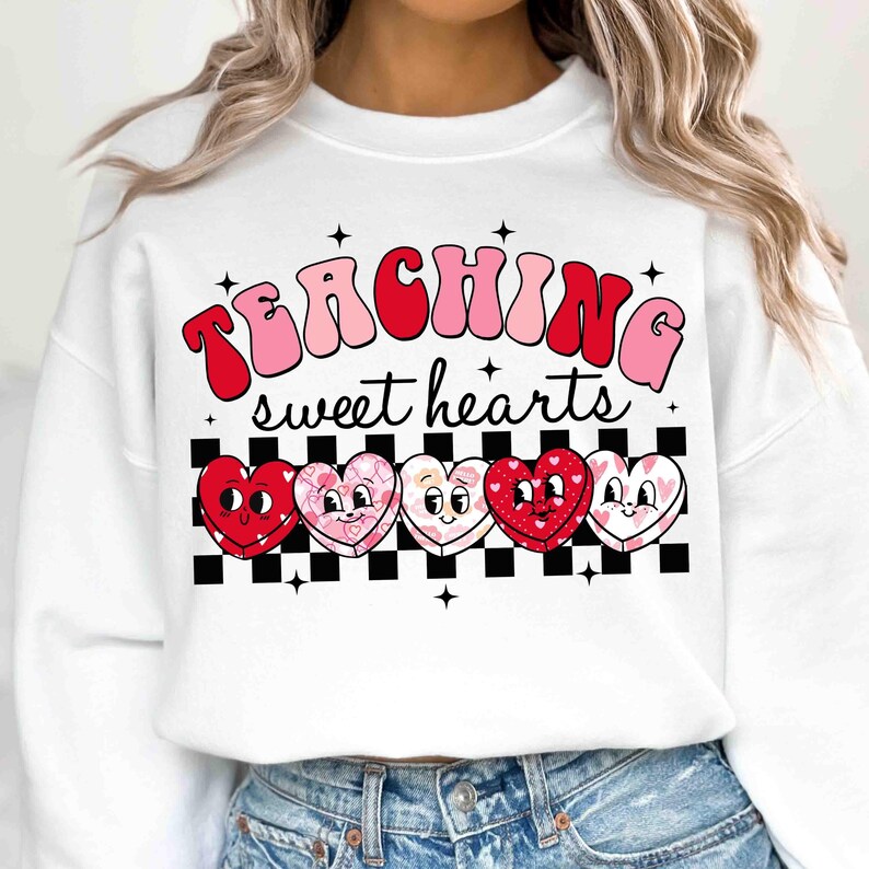Pu&ograve; includere: Felpa bianca con il testo "TEACHING sweet hearts" in lettere rosa e rosse. Cinque cuori di cartoni animati con facce sono allineati sotto il testo su uno sfondo a scacchi bianco e nero.