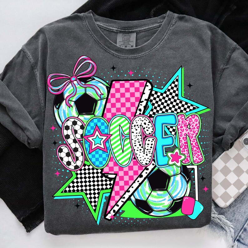 Puede incluir: Una camiseta gris oscuro con un dise&ntilde;o gr&aacute;fico de un bal&oacute;n de f&uacute;tbol, un rayo y la palabra "SOCCER" en letras estilizadas de colores. El dise&ntilde;o presenta un patr&oacute;n de cuadros rosa y blanco, estrellas blancas y negras y un patr&oacute;n de remolino verde y azul.