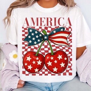 Puede incluir: Camiseta blanca con un dise&ntilde;o patri&oacute;tico. El gr&aacute;fico incluye dos cerezas rojas con estrellas blancas, un lazo con la bandera estadounidense y la palabra "AMERICA" en letras rojas. El fondo tiene un patr&oacute;n de cuadros rojos y blancos.