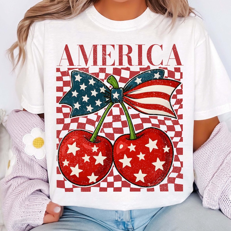 Puede incluir: Camiseta blanca con un dise&ntilde;o patri&oacute;tico. El gr&aacute;fico incluye dos cerezas rojas con estrellas blancas, un lazo con la bandera estadounidense y la palabra "AMERICA" en letras rojas. El fondo tiene un patr&oacute;n de cuadros rojos y blancos.
