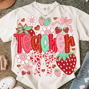 Op de afbeelding: Een wit T-shirt met een rood, roze en groen grafisch ontwerp met de tekst "Teacher" met aardbeien, bloemen en strikken.