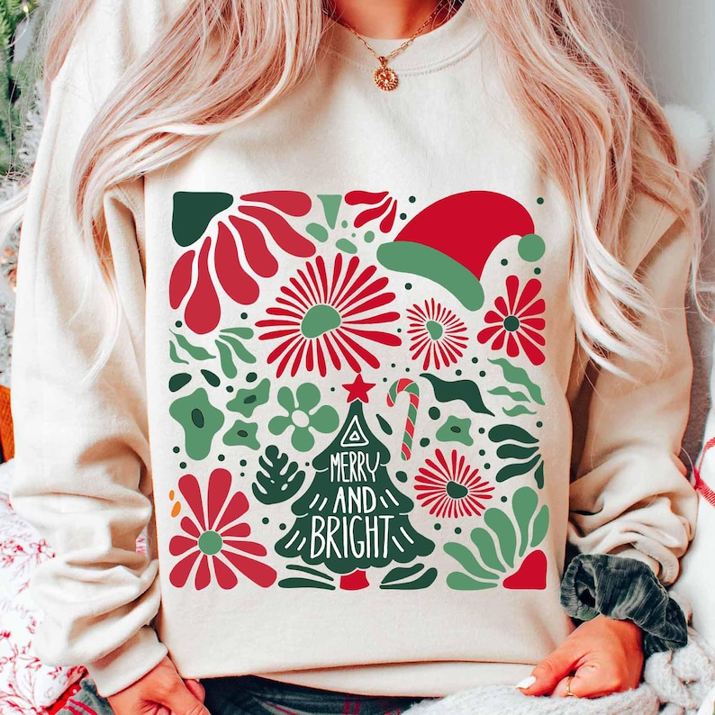 Puede incluir: Sudadera color crema con un dise&ntilde;o festivo. El gr&aacute;fico presenta flores rojas, hojas verdes, un gorro de Pap&aacute; Noel y un &aacute;rbol de Navidad con las palabras "MERRY AND BRIGHT". Tambi&eacute;n se incluye un bast&oacute;n de caramelo.