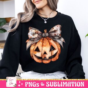 May include: A black sweatshirt with a watercolor illustration of a pumpkin with a bow. The pumpkin has a jack-o'-lantern face and is orange with brown accents. The bow is brown with white polka dots.