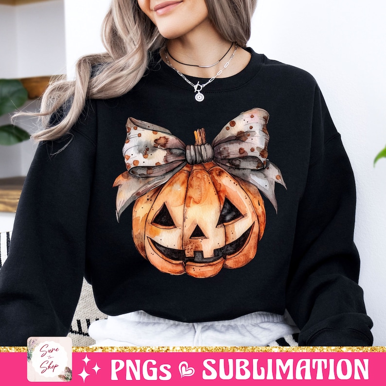 May include: A black sweatshirt with a watercolor illustration of a pumpkin with a bow. The pumpkin has a jack-o'-lantern face and is orange with brown accents. The bow is brown with white polka dots.