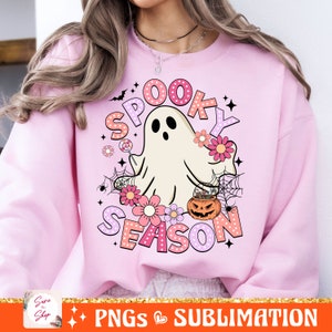 May include: Pink sweatshirt with a white ghost holding a lollipop and a pumpkin with a spiderweb design. The text "Spooky Season" is written in pink, orange, and white letters with polka dots.
