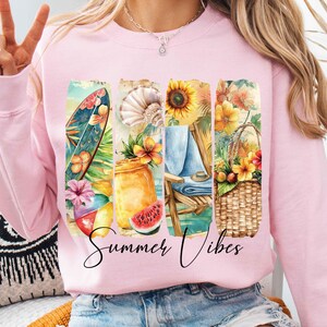 May include: Pink sweatshirt with a graphic design of a surfboard, seashells, sunflowers, a beach chair, and a basket of fruit. The text "Summer Vibes" is printed below the graphic.
