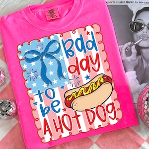 Puede incluir: Una camiseta rosa brillante con un gr&aacute;fico que dice "Bad Day to be a Hot Dog". El gr&aacute;fico incluye un lazo azul, fuegos artificiales, estrellas y un perrito caliente con mostaza. La camiseta es de la marca Comfort Colors.