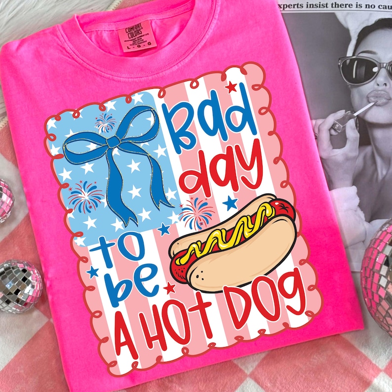 Puede incluir: Una camiseta rosa brillante con un gr&aacute;fico que dice "Bad Day to be a Hot Dog". El gr&aacute;fico incluye un lazo azul, fuegos artificiales, estrellas y un perrito caliente con mostaza. La camiseta es de la marca Comfort Colors.
