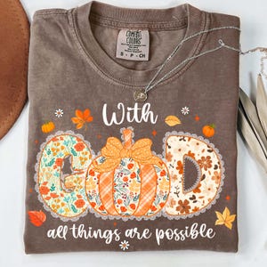 May include: Brown t-shirt with the words "With God all things are possible." The word "GOD" is designed with a pumpkin in the center and floral patterns. A silver necklace is draped across the shirt.