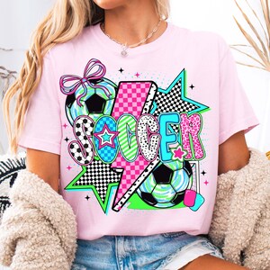 Puede incluir: Camiseta rosa con un dise&ntilde;o gr&aacute;fico que presenta la palabra "SOCCER" en letras de colores, un rayo, estrellas y balones de f&uacute;tbol.