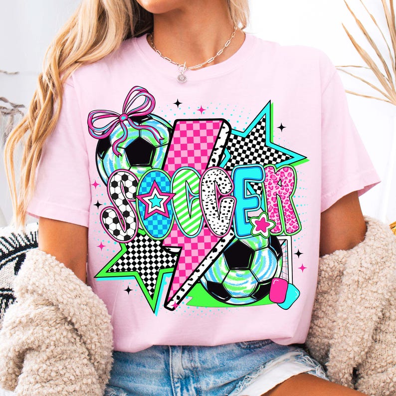 Puede incluir: Camiseta rosa con un dise&ntilde;o gr&aacute;fico que presenta la palabra "SOCCER" en letras de colores, un rayo, estrellas y balones de f&uacute;tbol.