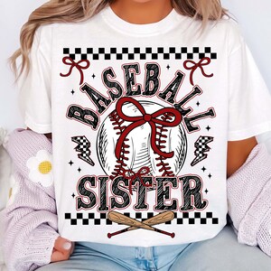 May include: White t-shirt with a baseball-themed graphic. The design features a baseball with a red bow, the words "BASEBALL SISTER" in a decorative font, and crossed baseball bats at the bottom. Black and red accents complete the design.