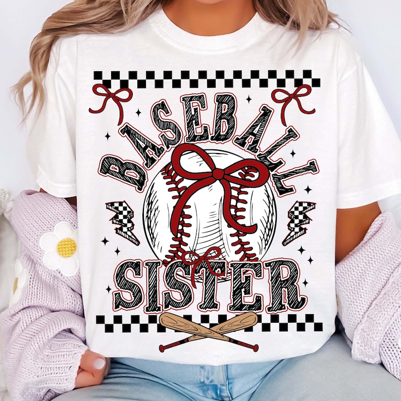 May include: White t-shirt with a baseball-themed graphic. The design features a baseball with a red bow, the words "BASEBALL SISTER" in a decorative font, and crossed baseball bats at the bottom. Black and red accents complete the design.