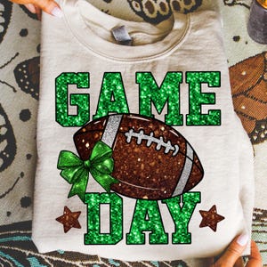Puede incluir: Sudadera crema con dise&ntilde;o de f&uacute;tbol americano. Las palabras "GAME DAY" est&aacute;n en letras verdes brillantes, con un bal&oacute;n de f&uacute;tbol marr&oacute;n en el centro. Un lazo verde y dos estrellas marrones completan el dise&ntilde;o.