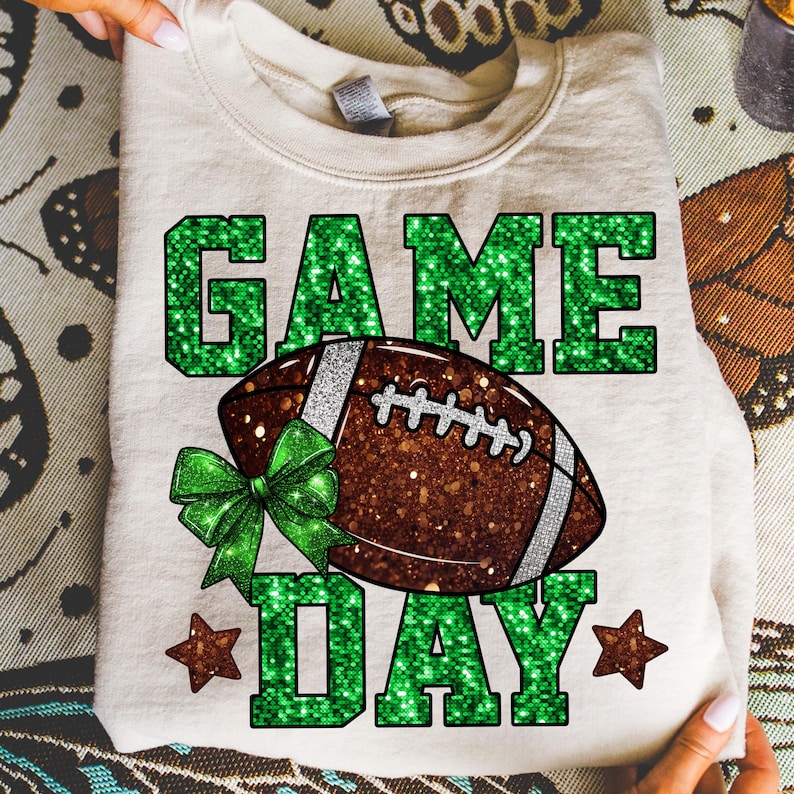 Puede incluir: Sudadera crema con dise&ntilde;o de f&uacute;tbol americano. Las palabras "GAME DAY" est&aacute;n en letras verdes brillantes, con un bal&oacute;n de f&uacute;tbol marr&oacute;n en el centro. Un lazo verde y dos estrellas marrones completan el dise&ntilde;o.
