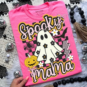 Puede incluir: Camiseta rosa brillante con un dise&ntilde;o de Halloween. El dise&ntilde;o presenta las palabras "Spooky Mama", un fantasma, una calabaza, una ara&ntilde;a y un murci&eacute;lago. La camiseta tiene cuello redondo y mangas cortas.